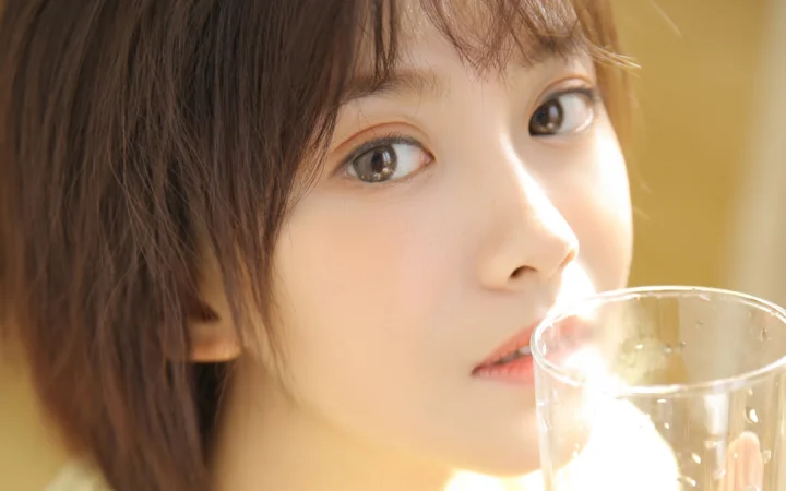 一位女性在暖色光线下手持透明玻璃杯，面部特写占据画面大部分。