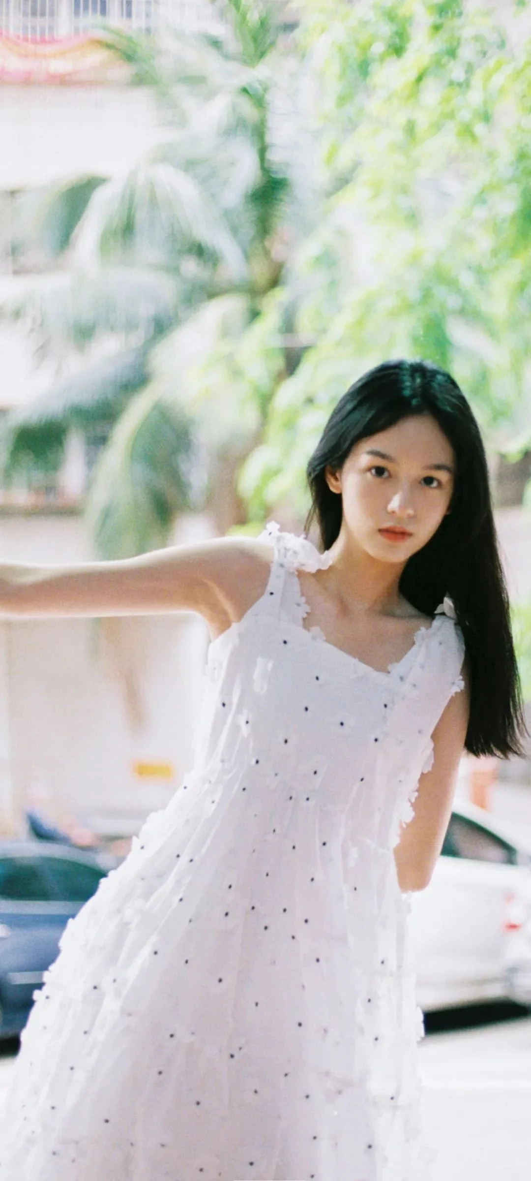긴 머리의 여성이 흰색 원피스를 입고 초록 나무와 거리 풍경을 배경으로 서 있습니다.