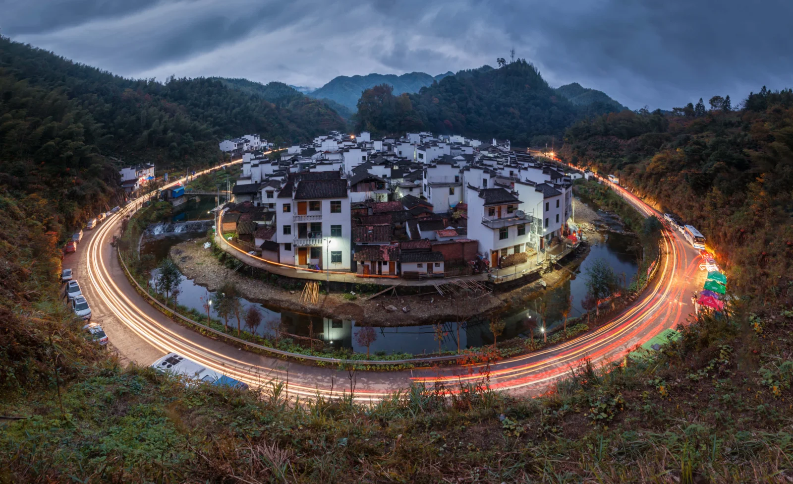 群山环抱的白墙黑瓦村落被环形道路与绚丽车流光轨环绕。