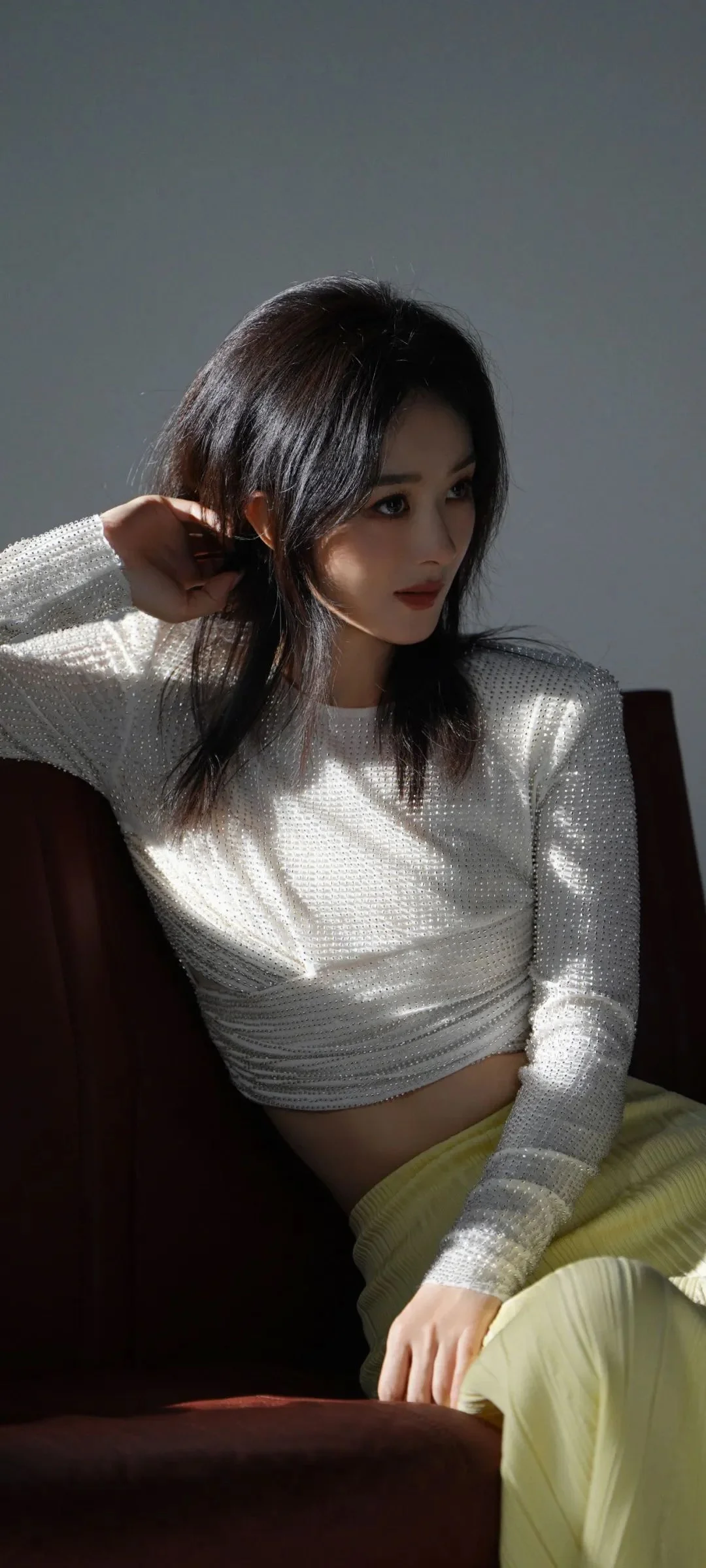 一名成年女子坐在椅子上，穿着白色长袖上衣和浅黄色下装，沐浴在室内光线中。
