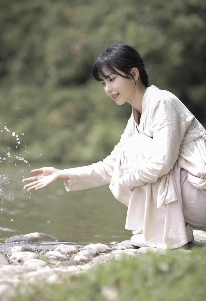 밝은 색 옷을 입은 여성이 개울가에 쪼그려 앉아 튀는 물방울에 손을 뻗고 있으며 뒤에는 초점이 흐린 초록 숲이 보입니다.