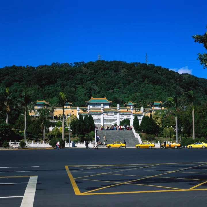 一座中式建筑坐落在郁郁葱葱的山坡前，前方道路上停着几辆黄色出租车。