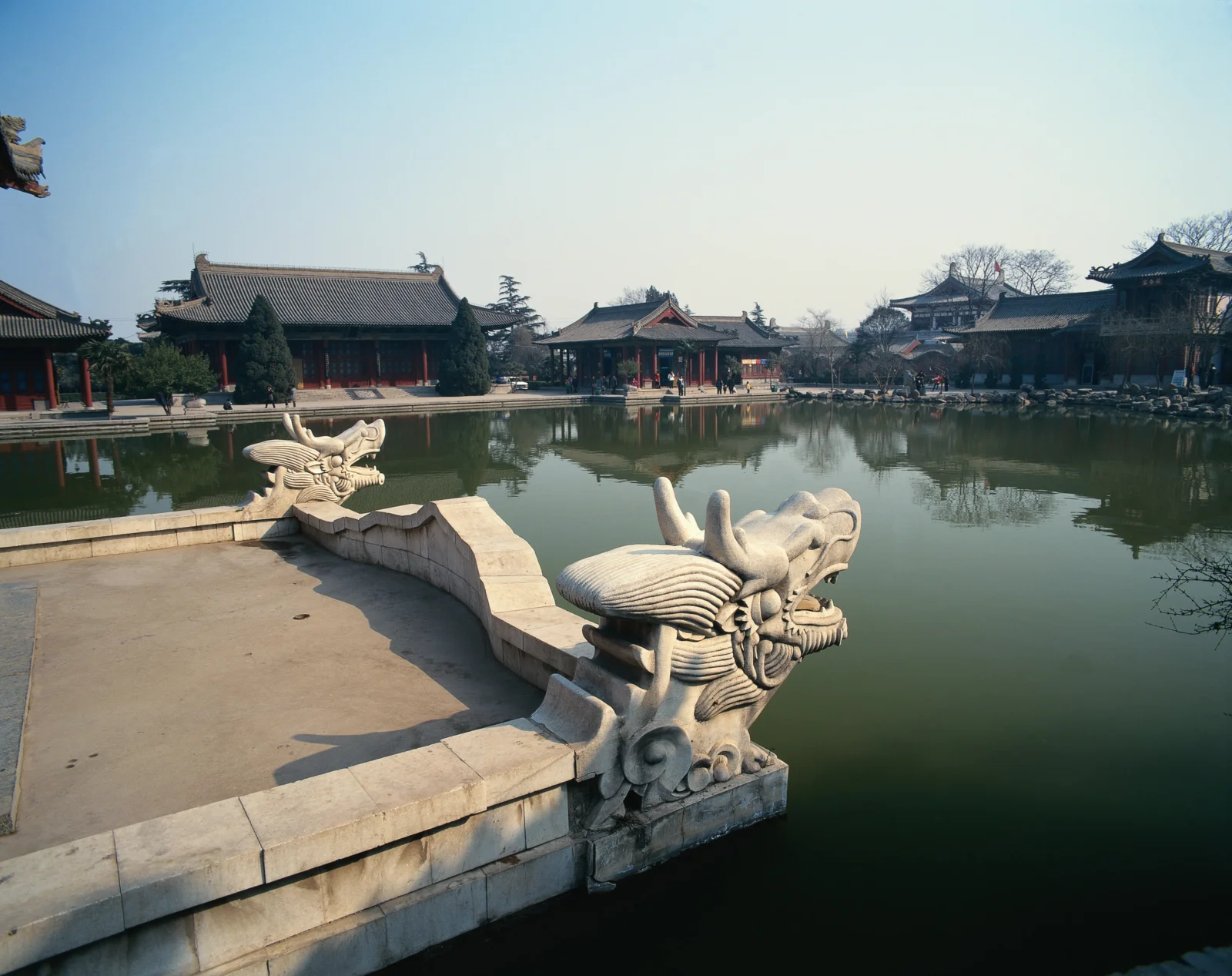 古建築群環繞著寧靜的池塘，前景可見雕刻精美的石質龍首欄杆。