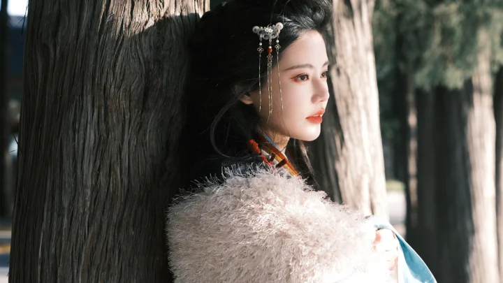 一名女性靠在树旁，以侧脸望向右侧，阳光穿过树木照亮她的面庞。