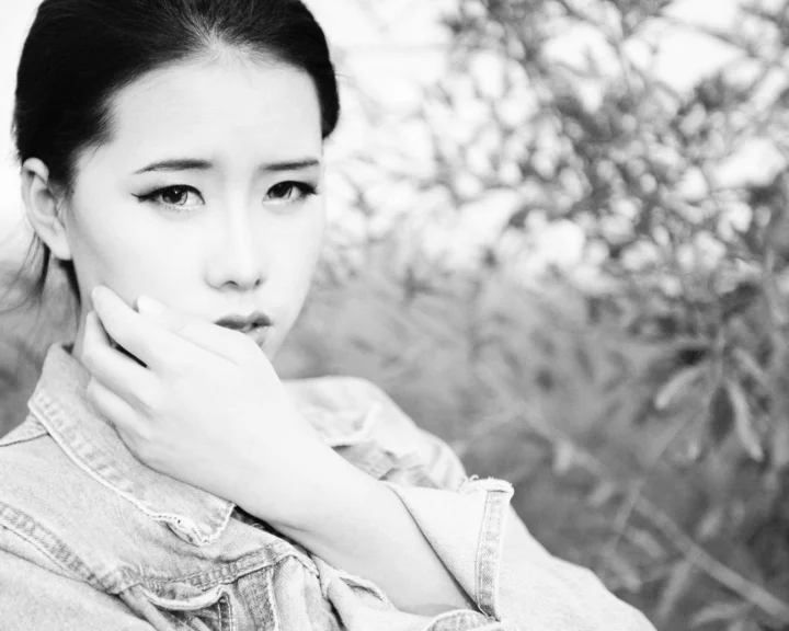 一名成年女性身穿牛仔外套，在户外树叶背景前以黑白风格入镜。