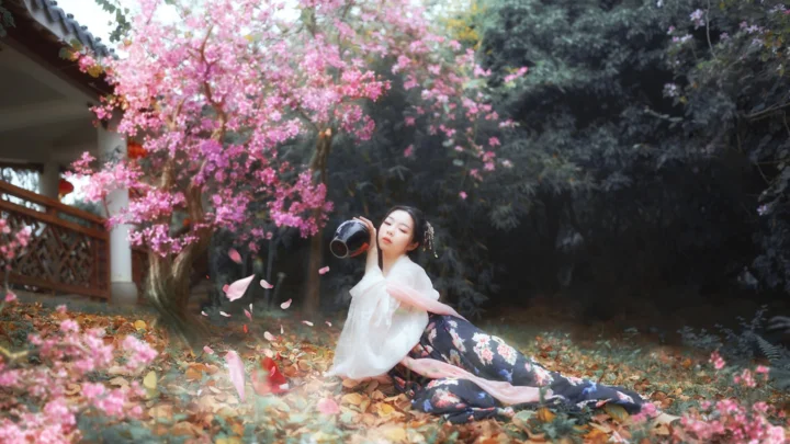 一名女子坐在粉色花树下的庭院落叶中。