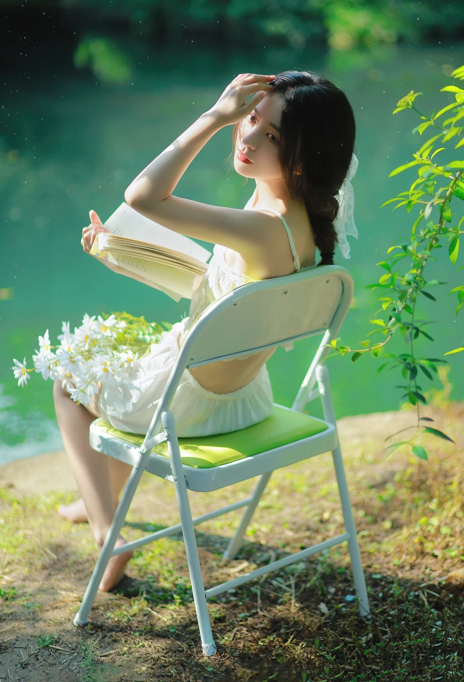 女性が湖畔の折りたたみ椅子に座り、本を手に白い花と緑の葉に囲まれている。
