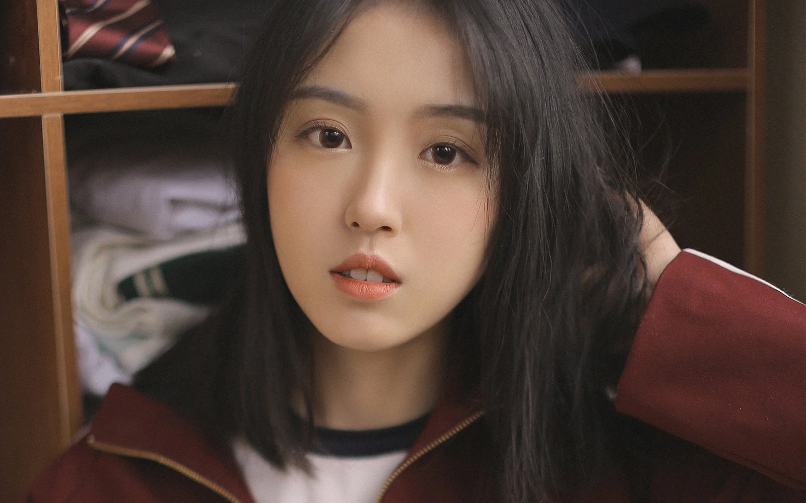긴 머리의 여성이 빨간 재킷을 입고 나무 옷장 앞에서 카메라를 바라보고 있습니다.