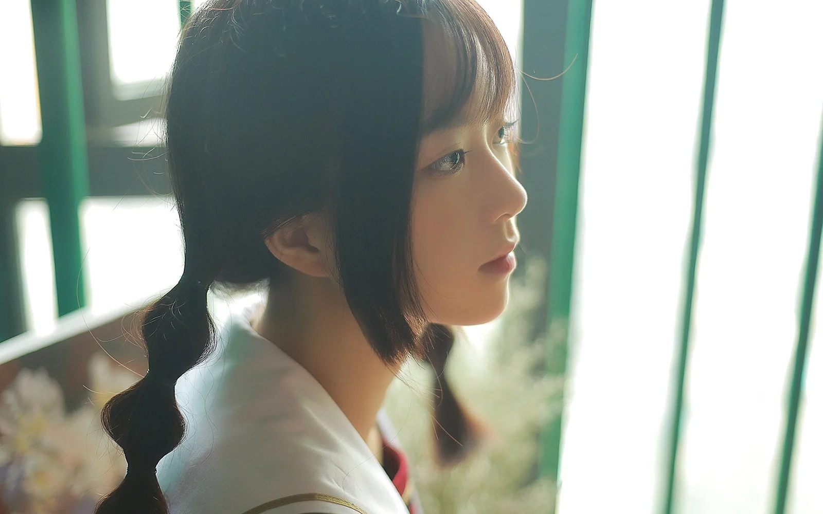 밝은 창가에서 땋은 머리와 흰색 상의를 한 여성이 옆모습을 보이고 있다.