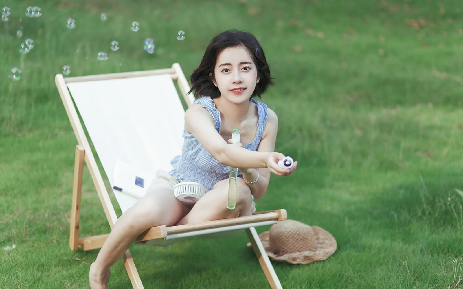 一名女子坐在绿草地上的躺椅上，手中拿着小物件，周围飘着泡泡。
