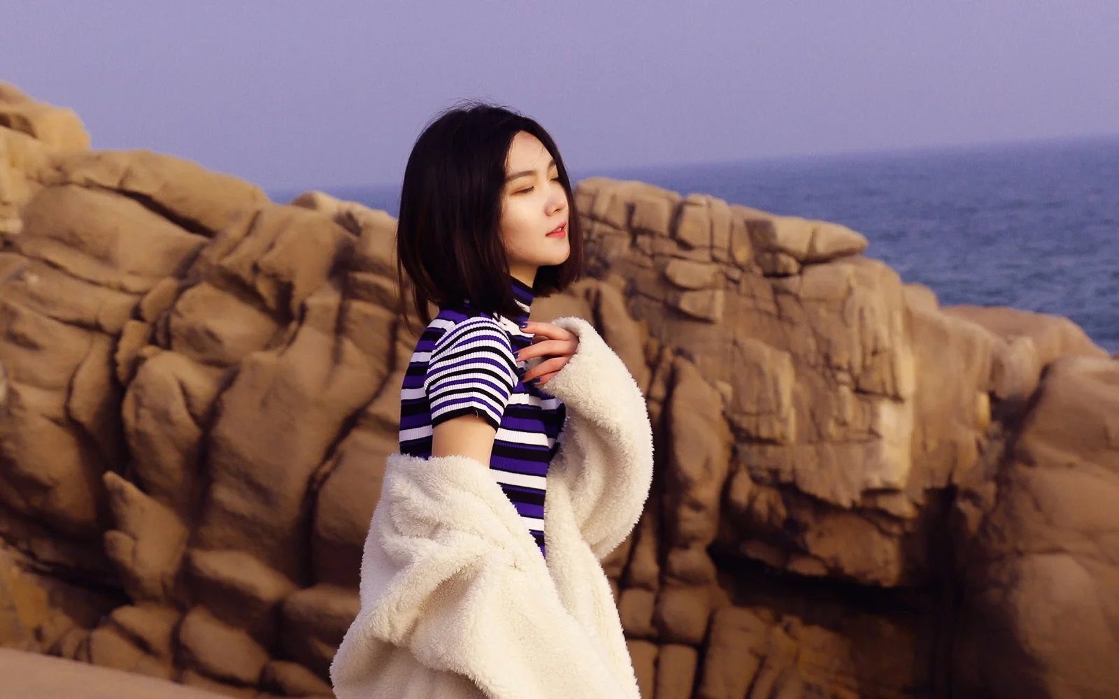 성인 여성이 해안 바위 앞에 서 있고 뒤로 탁 트인 바다가 보입니다.