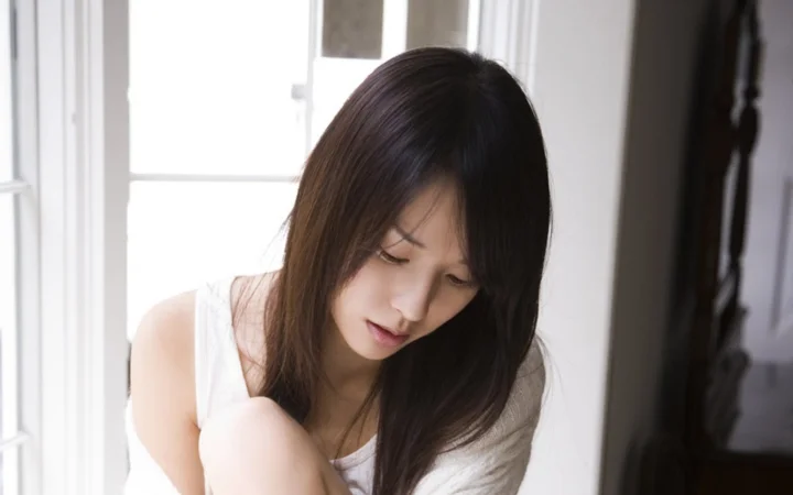 長い髪の女性が窓辺に座り、柔らかな室内光の中でうつむいています。