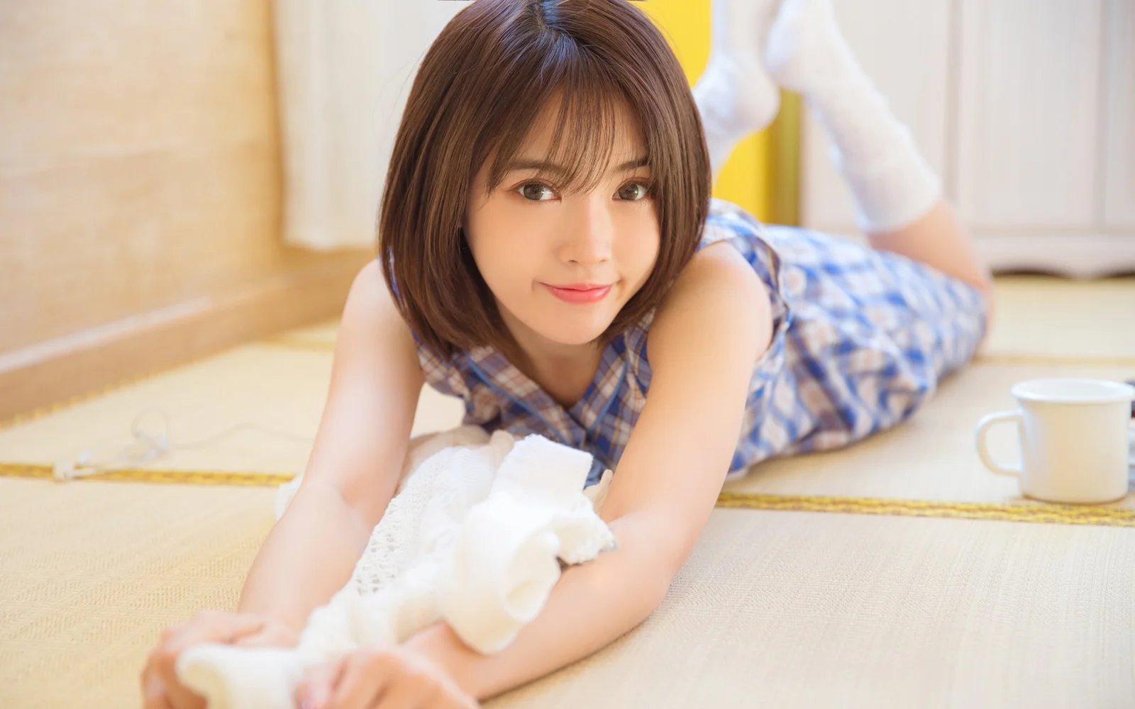 青いチェック柄のワンピースを着た笑顔の成人女性が、白いカップの隣で明るい床に横たわっています。