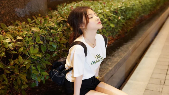 한 여성이 생울타리 옆 보도에 앉아 눈을 감고 자연광을 받고 있다.