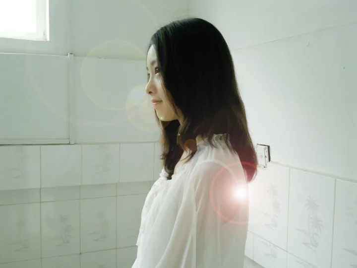 一名成年女性身穿白色上衣站在室内瓷砖墙前，侧脸面对左侧，画面带有明亮的镜头光晕。