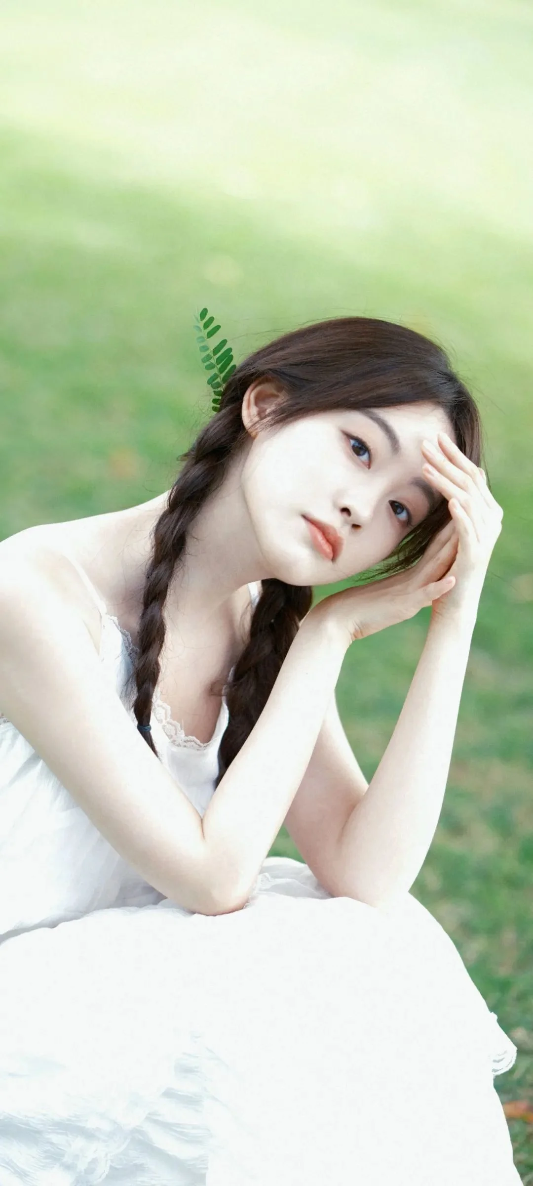 一位成年女性身穿白裙，在绿色户外背景前侧头托腮。