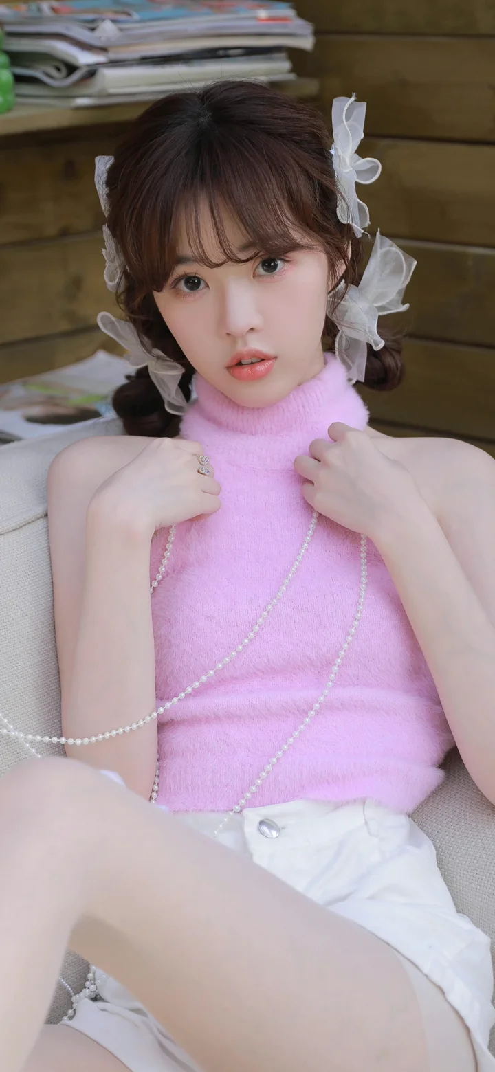 一名女子坐在室内椅子上，穿着粉色高领无袖针织衫，手持珍珠项链。