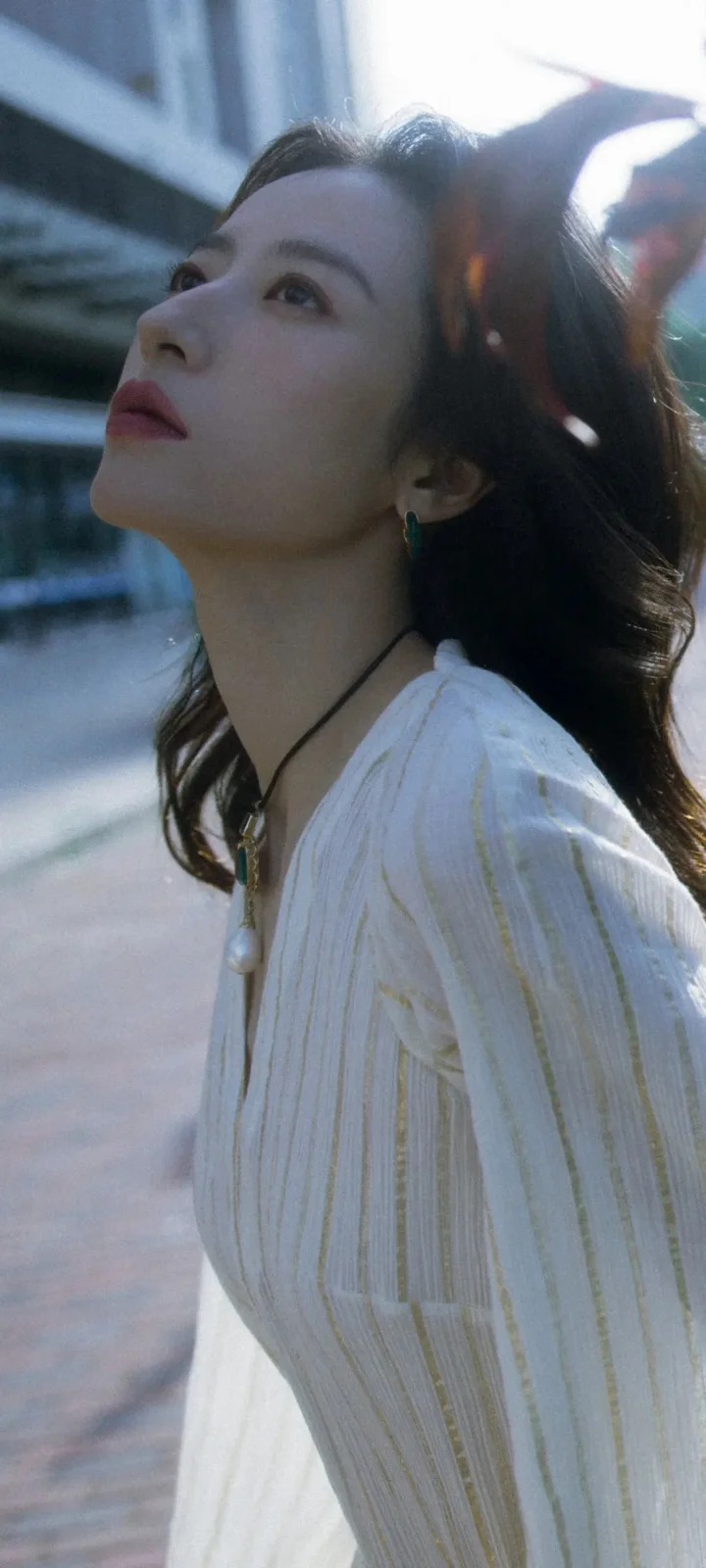 흰색 블라우스를 입은 성인 여성이 도시 건물과 거리를 배경으로 야외 햇빛 아래 서 있다.