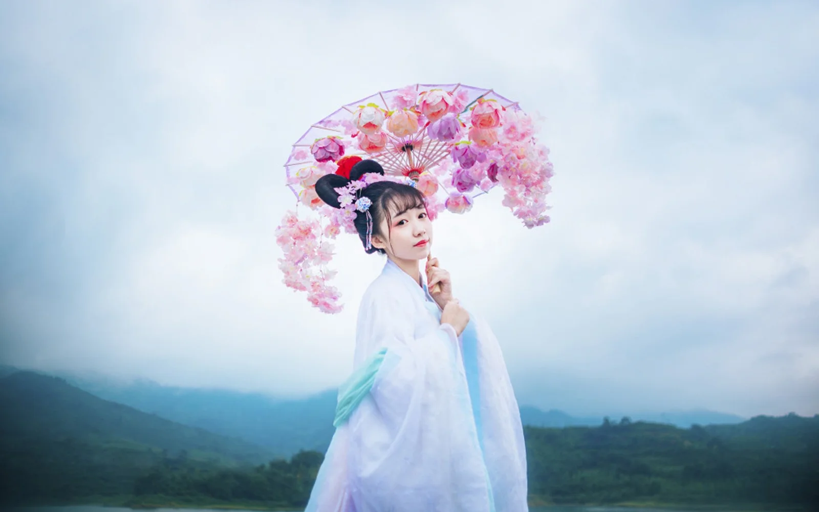 一名女子手持粉色花伞站在朦胧山景前。