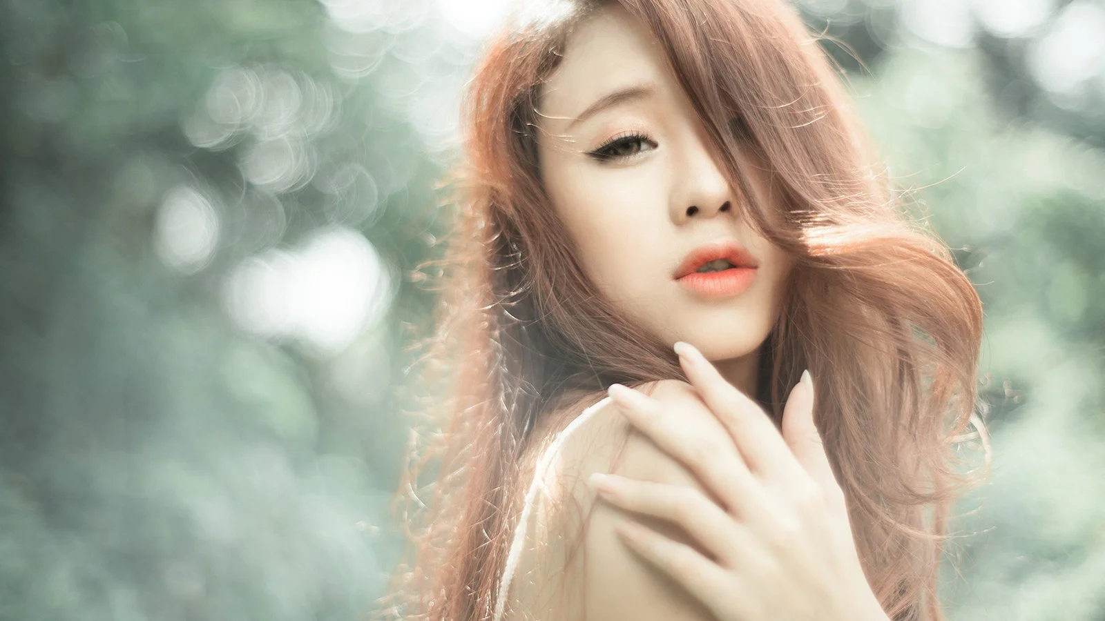 一位长发女性在柔和的户外散景背景前注视镜头，手靠近脸部。
