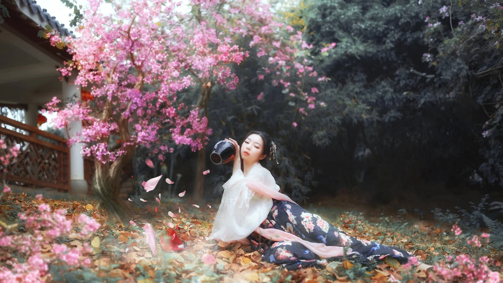 一名女子坐在粉色花树下的庭院落叶中。