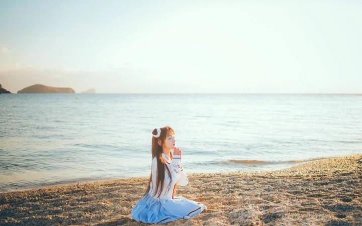 一名女子坐在海边的卵石沙滩上，面向平静的海面。