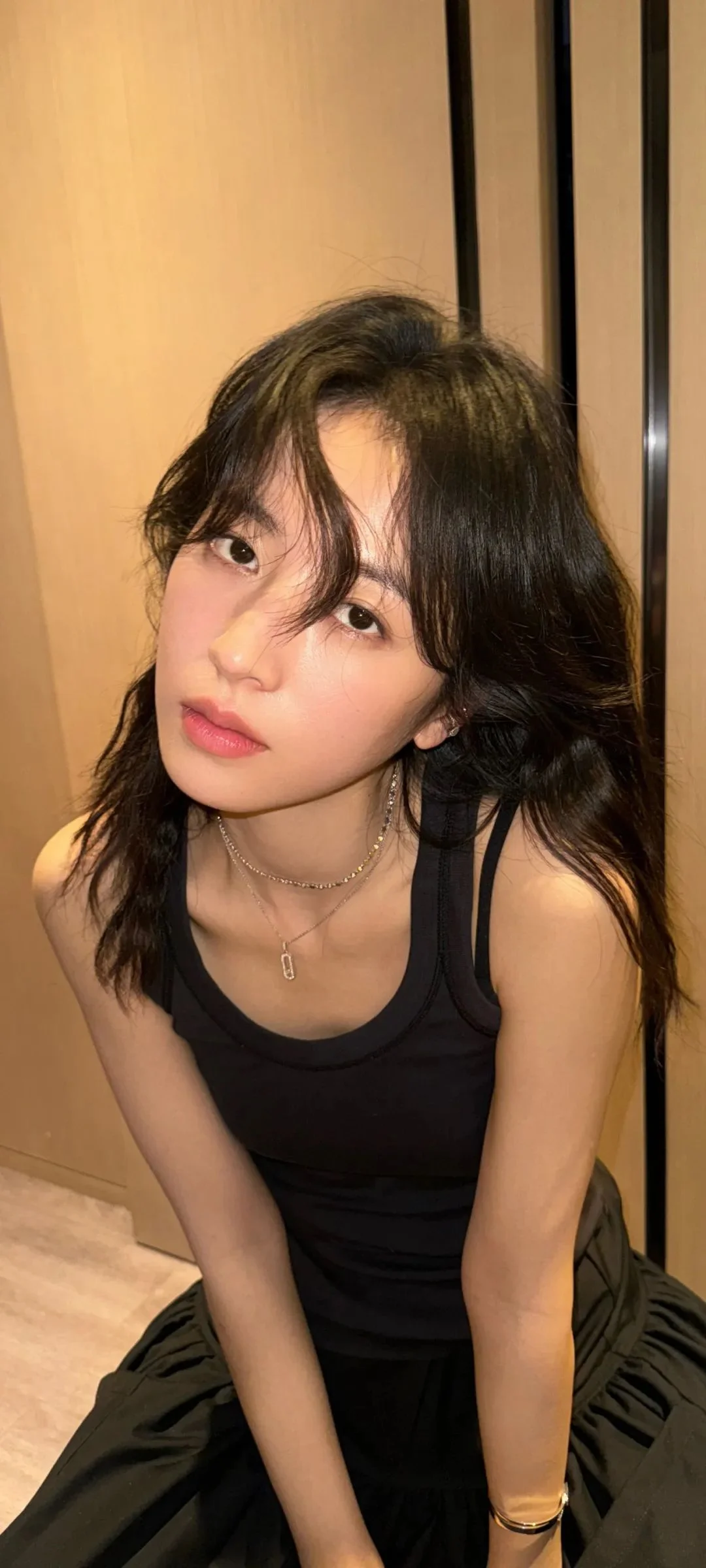 긴 머리의 여성이 검은색 민소매 상의를 입고 실내 나무 배경 앞에 앉아 있다.
