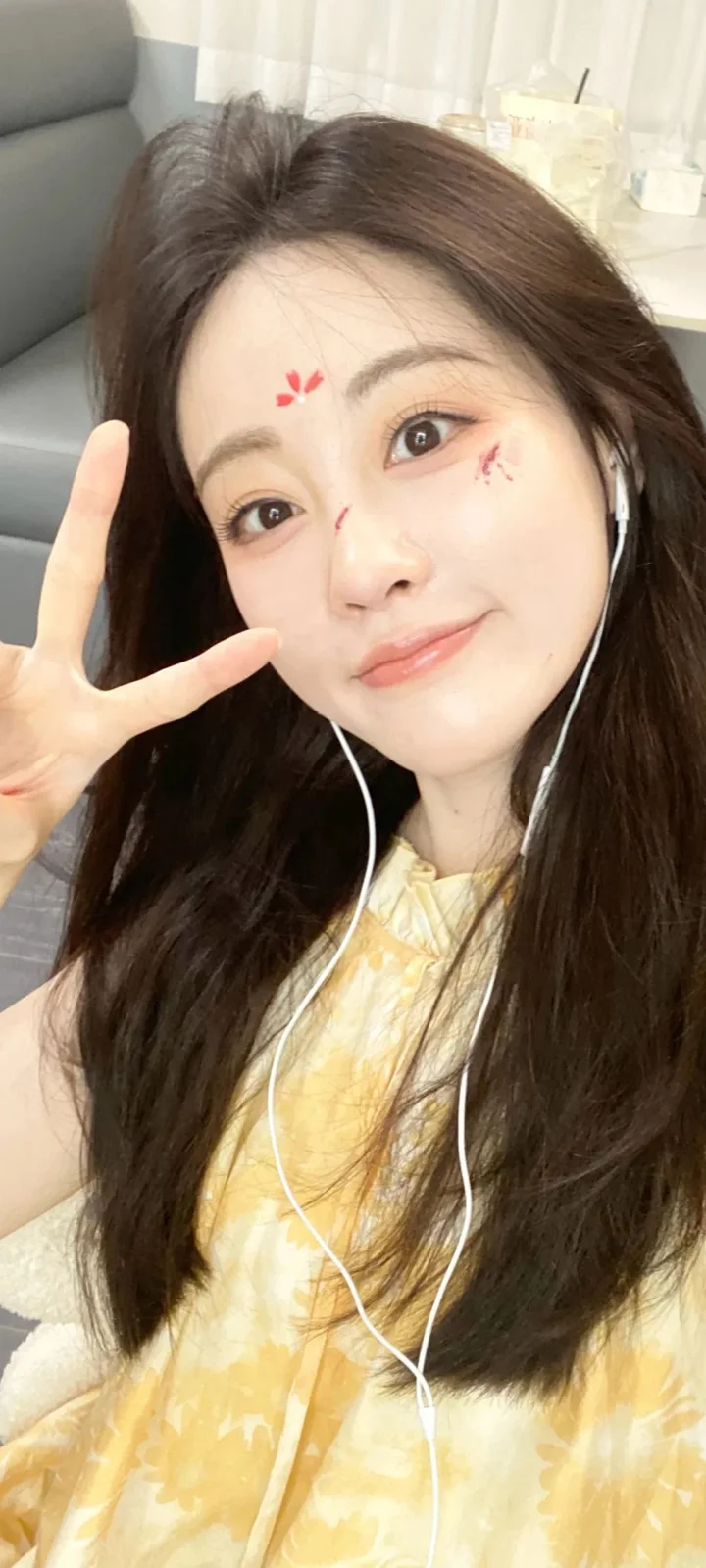 一名长发女性在室内对着镜头微笑自拍，手指比出手势并戴着有线耳机。
