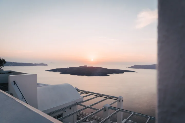 夕阳沉入平静的海面，远处是火山岛，前景可见圣托里尼的白色建筑。