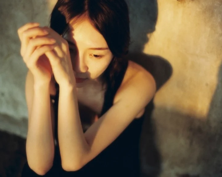 한 여성이 따뜻한 빛 아래 벽에 기대어 고개를 숙이고 있으며 얼굴과 팔에 빛과 그림자가 드리워져 있습니다.