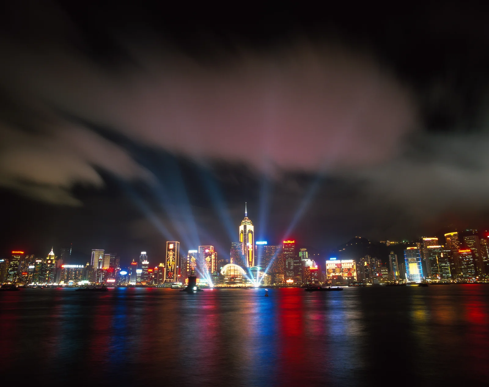 夜色中的城市天际线倒映在水面上，建筑灯光与探照灯照亮了天空。