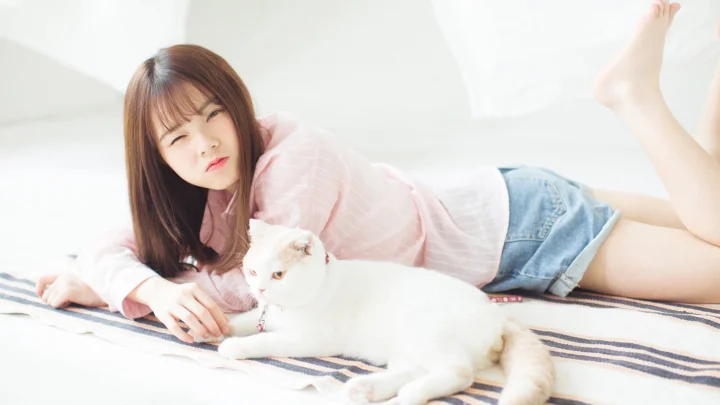 一名成年女子穿着粉色上衣和牛仔短裤躺在床上，身旁有一只白猫。