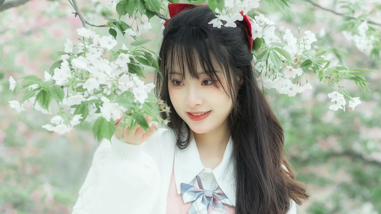 긴 검은 머리의 여성이 흰 스웨터와 나비넥타이를 착용하고 활짝 핀 흰 꽃 사이에 서 있습니다.