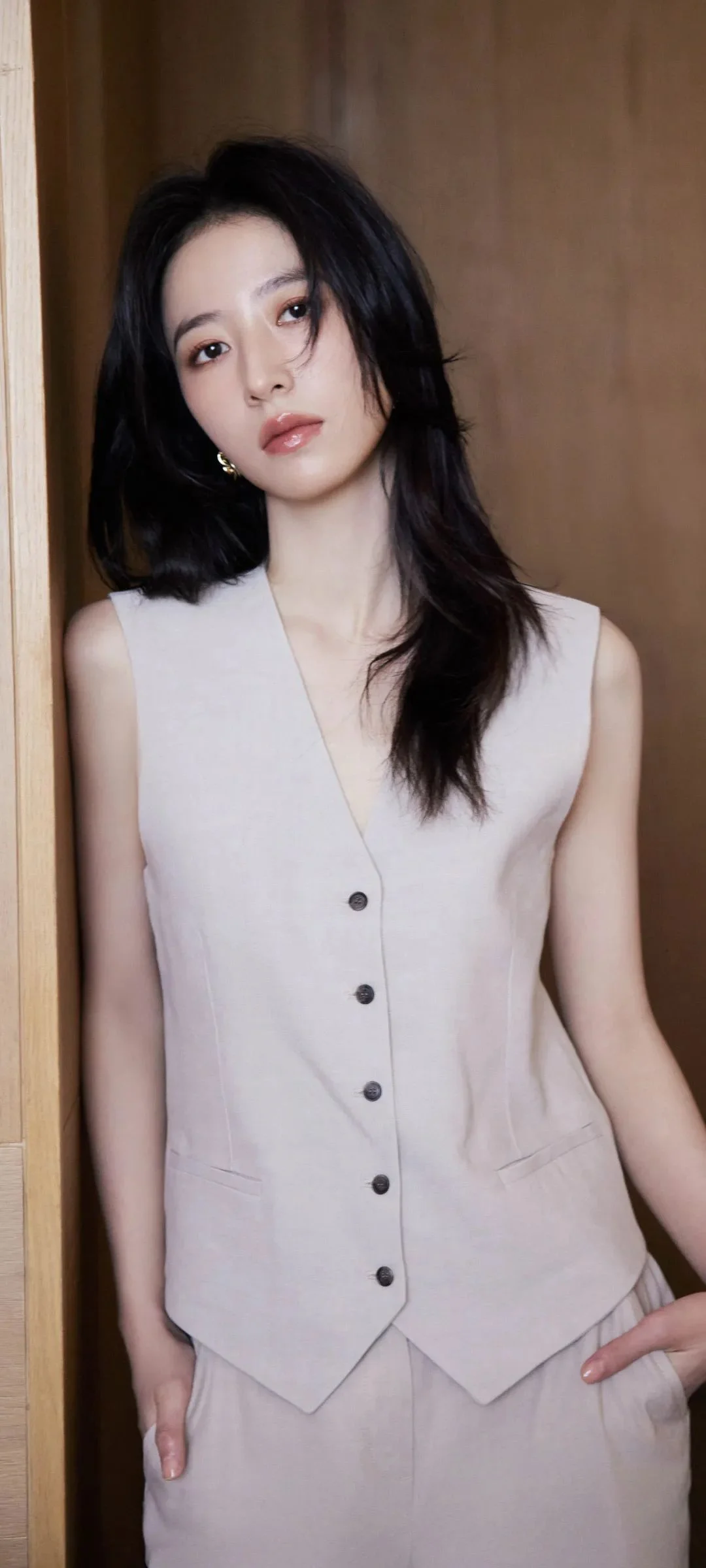 一名成年女性身穿浅色无袖马甲站在木质室内背景前。