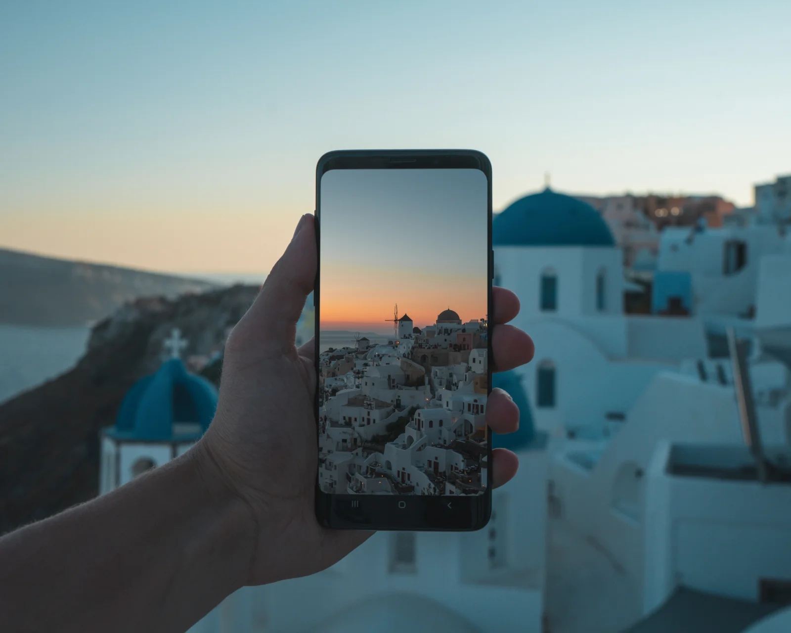手に持ったスマートフォンの画面に、夕暮れ時のサントリーニ島の白い建物と青いドームの教会が映っている。