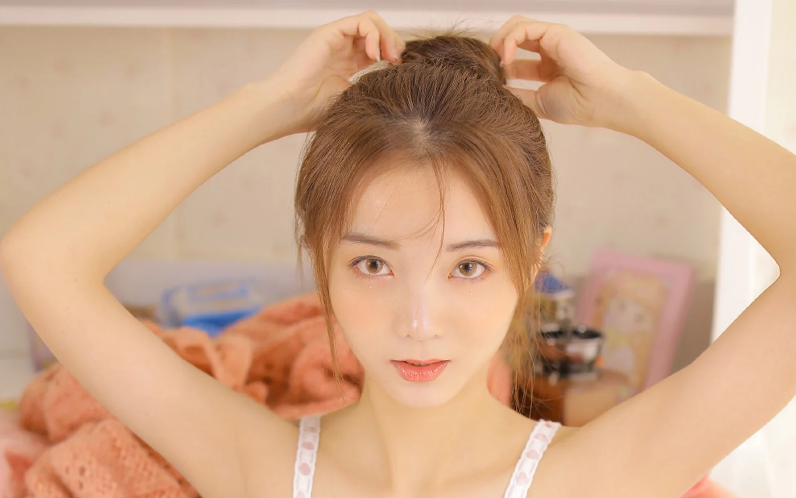 成人の女性が室内で両腕を上げて髪を整えながら、正面からカメラを見つめています。