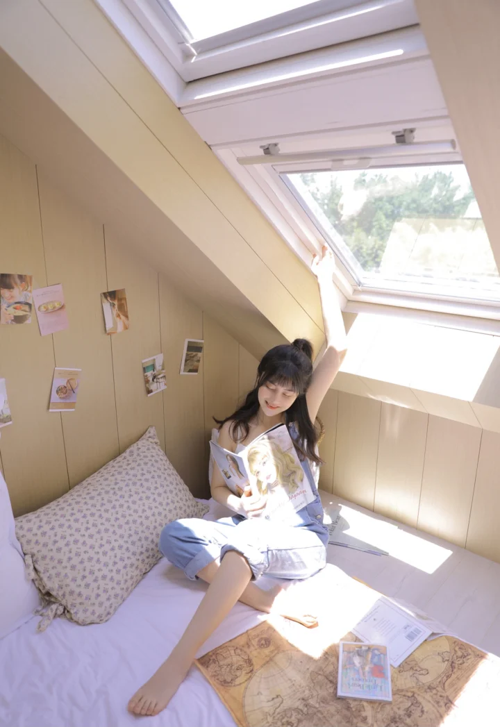 한 여성이 침실 침대에 앉아 채광창으로 들어오는 햇빛 속에서 잡지를 읽고 있다.