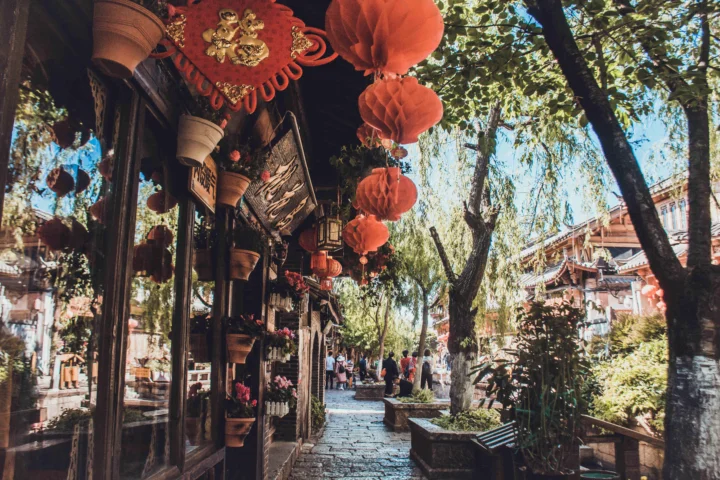 一条铺着石板的古镇街巷两旁分布着传统建筑、树木和悬挂的红灯笼，远处可见行人。