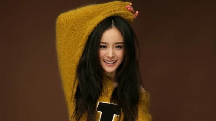 一名长发女性穿着黄色针织衫，在棕色背景前微笑摆姿势。