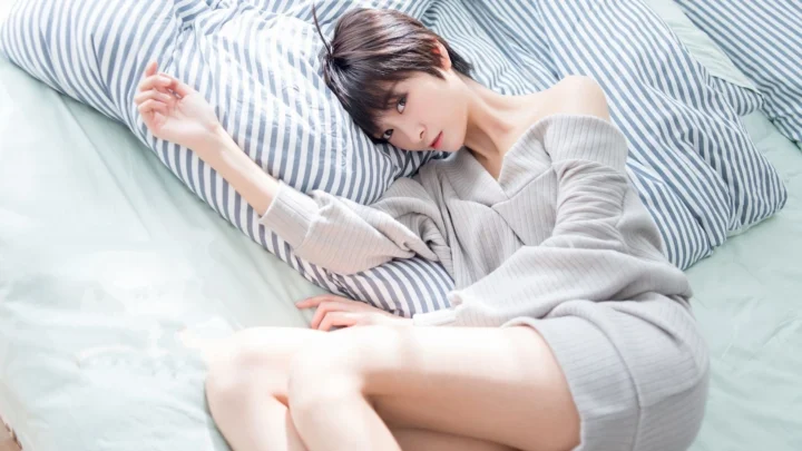 연한 색 니트를 입은 성인 여성이 줄무늬 베개에 머리를 기대고 침대에 누워 있습니다.