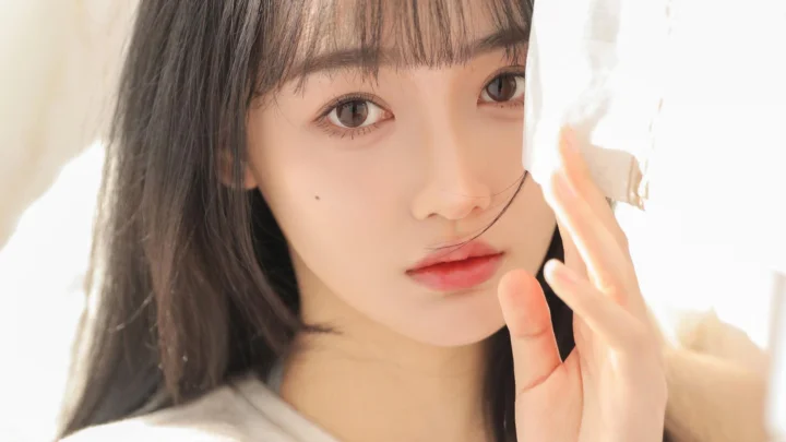 一位女性在柔和明亮的光线下靠近镜头，手指轻触面部一侧。