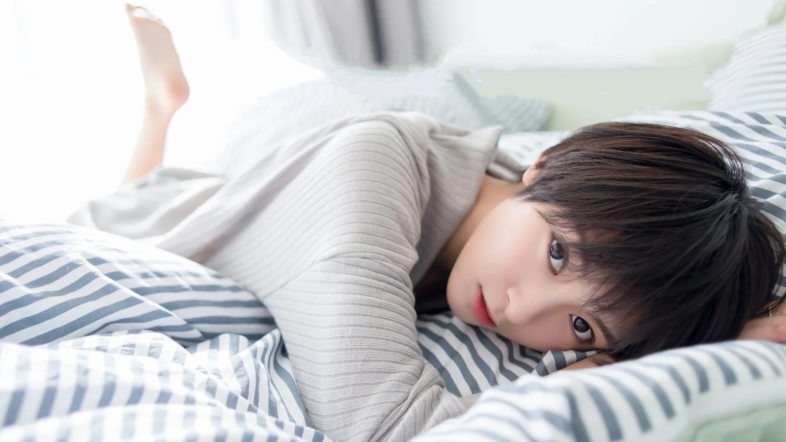 연한 회색 니트를 입은 인물이 줄무늬 침구 위에 엎드려 있습니다.