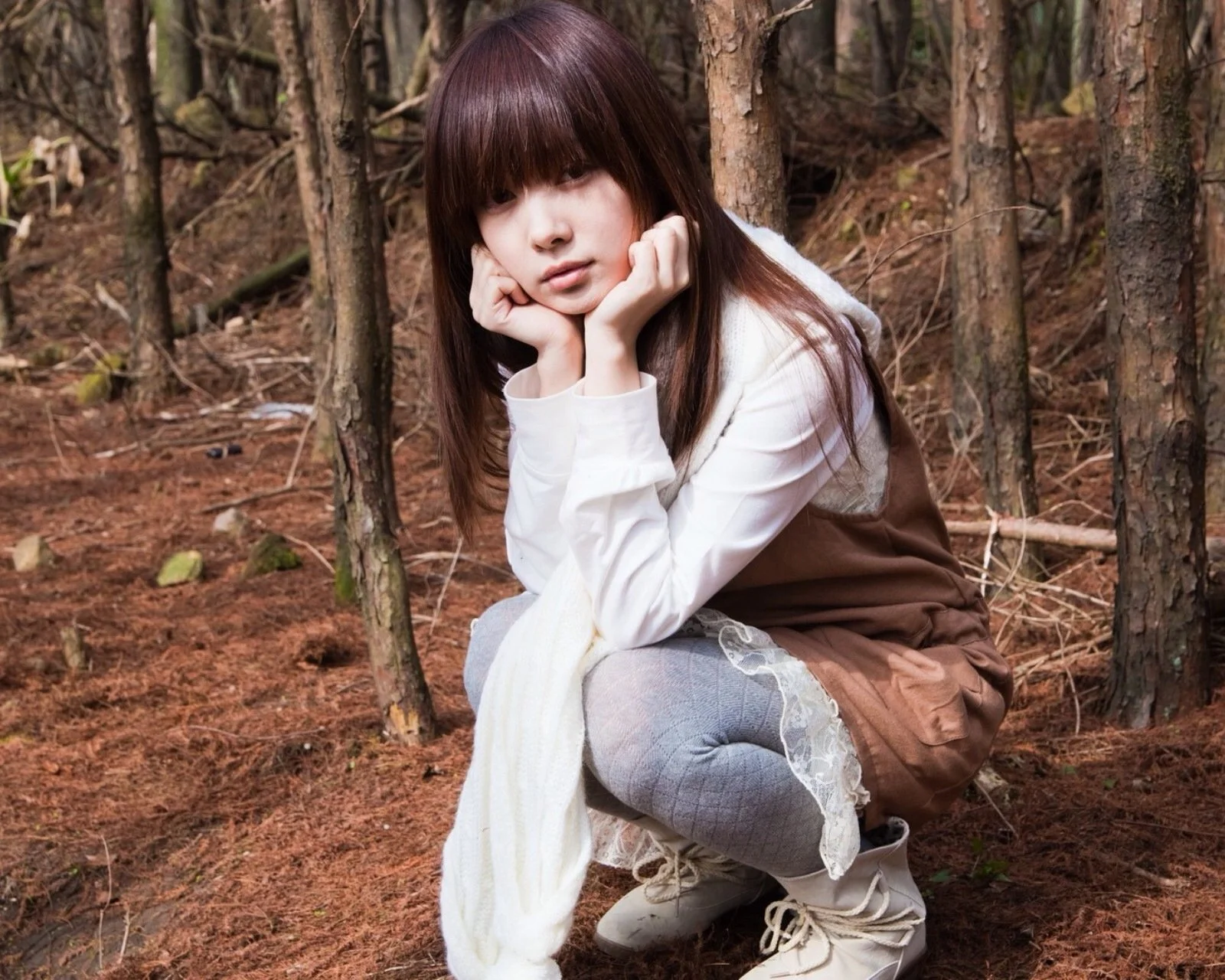 一名长发女子坐在森林地面上，双手托腮望向镜头。