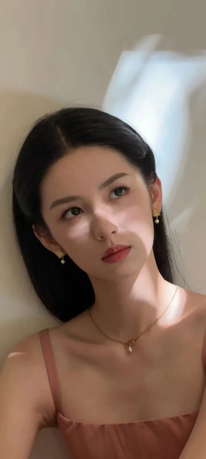 一名女性靠在室内墙边，在柔和光影下看向镜头。