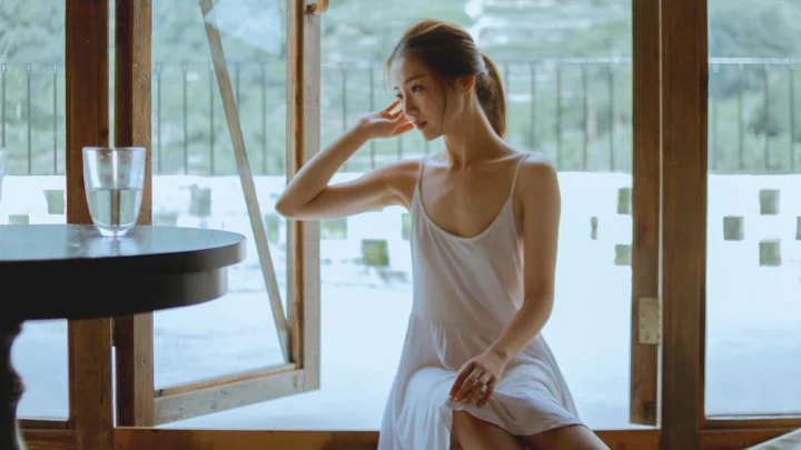 一名身穿白色連衣裙的女子坐在木質窗邊，窗外映入明亮的自然光。