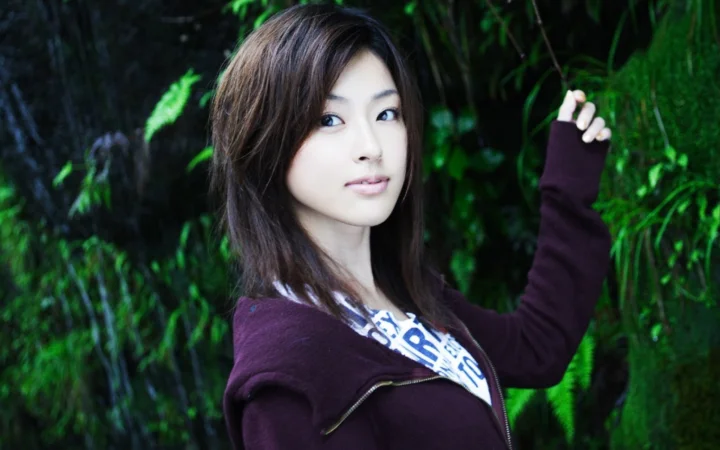 짙은 긴 머리의 여성이 초록색 숲을 배경으로 서서 짙은 보라색 재킷을 입고 카메라를 바라보고 있습니다.