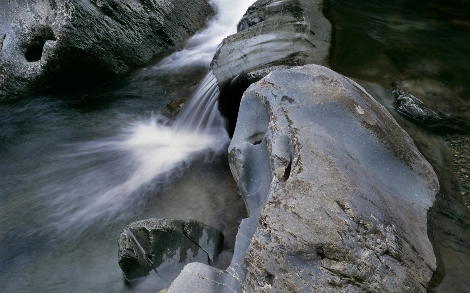 溪水在深色岩石之间形成小瀑布并流入浅滩。