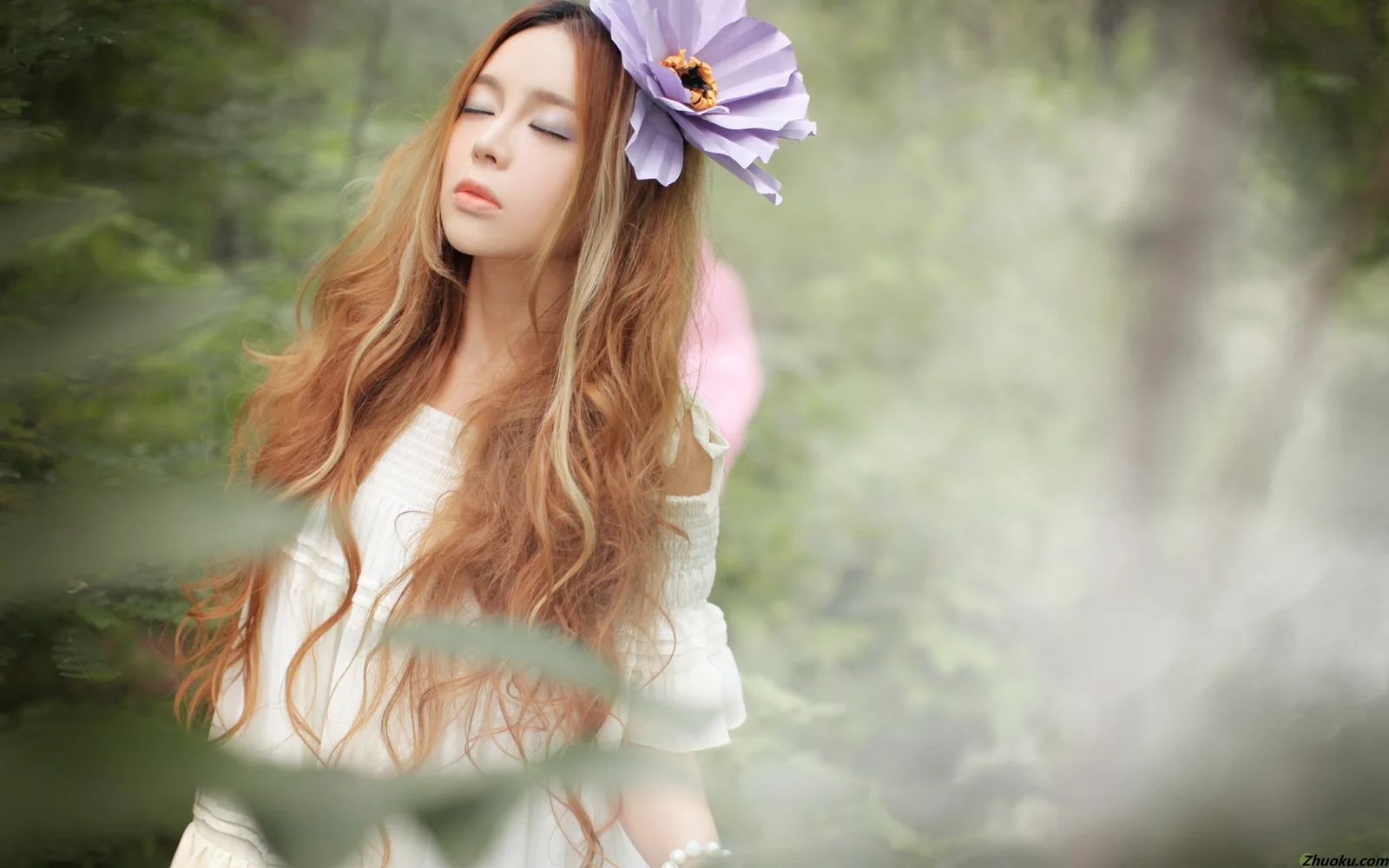 보라색 꽃 장식을 한 긴 머리 여성이 흐릿한 숲을 배경으로 서 있다.