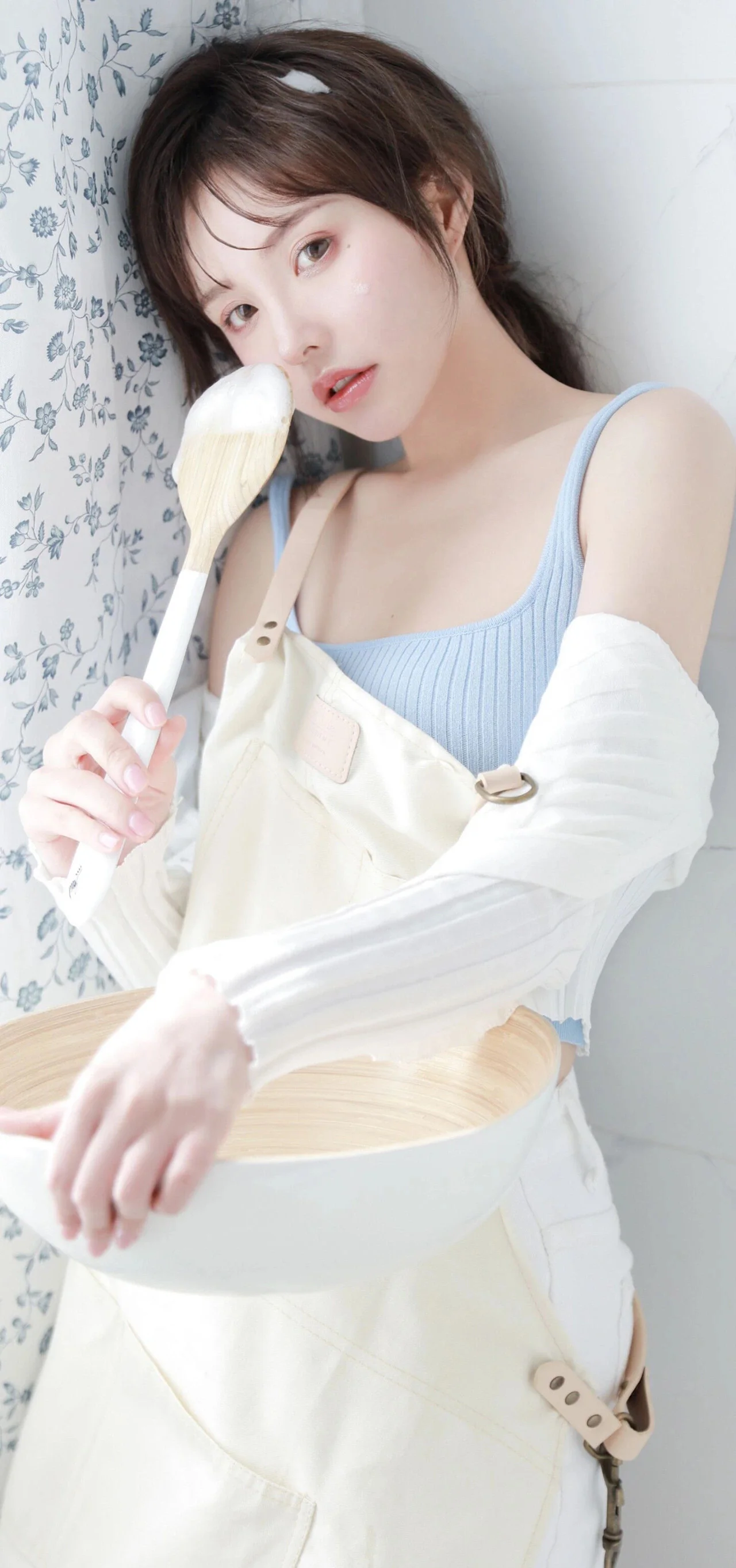淡い青色のトップスと白いエプロンを着た成人女性が、明るい室内でボウルとヘラを持っている。