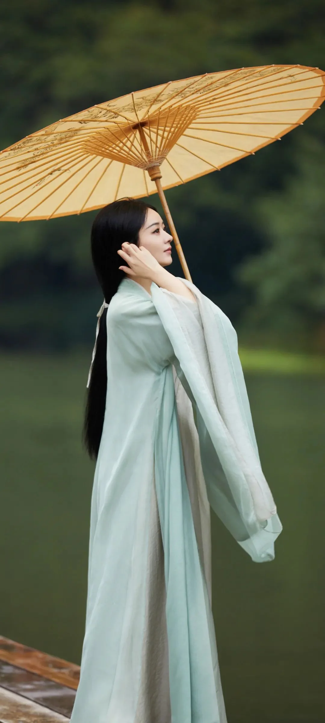 밝은 전통 의상을 입은 성인 여성이 초록빛 배경이 흐릿한 야외에서 종이 우산을 들고 있다.